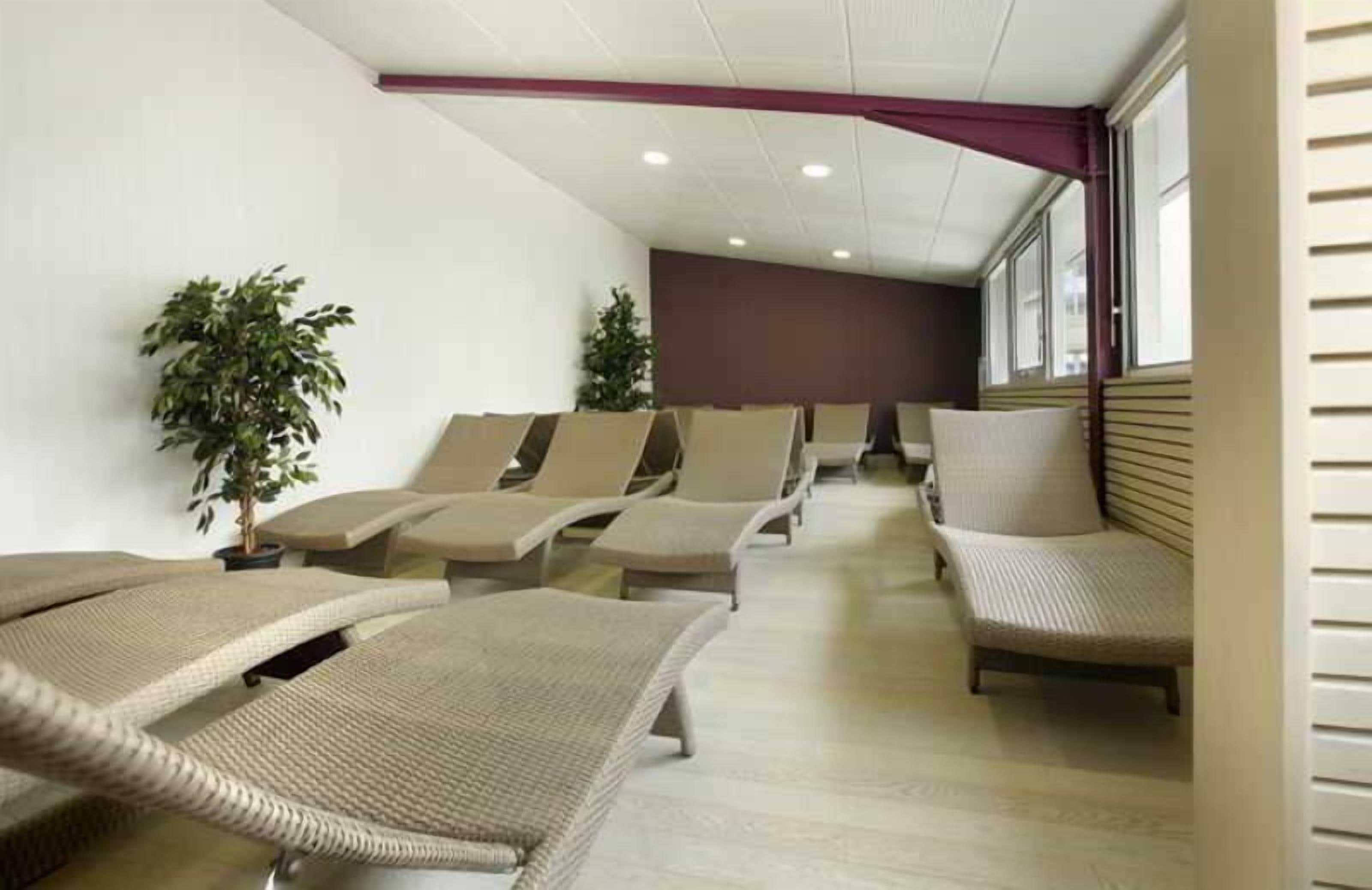 Ein Zimmer mit mehreren beigefarbenen Loungesesseln, die in Reihen angeordnet sind, einer Zimmerpflanze und einem Fenster mit Jalousien.