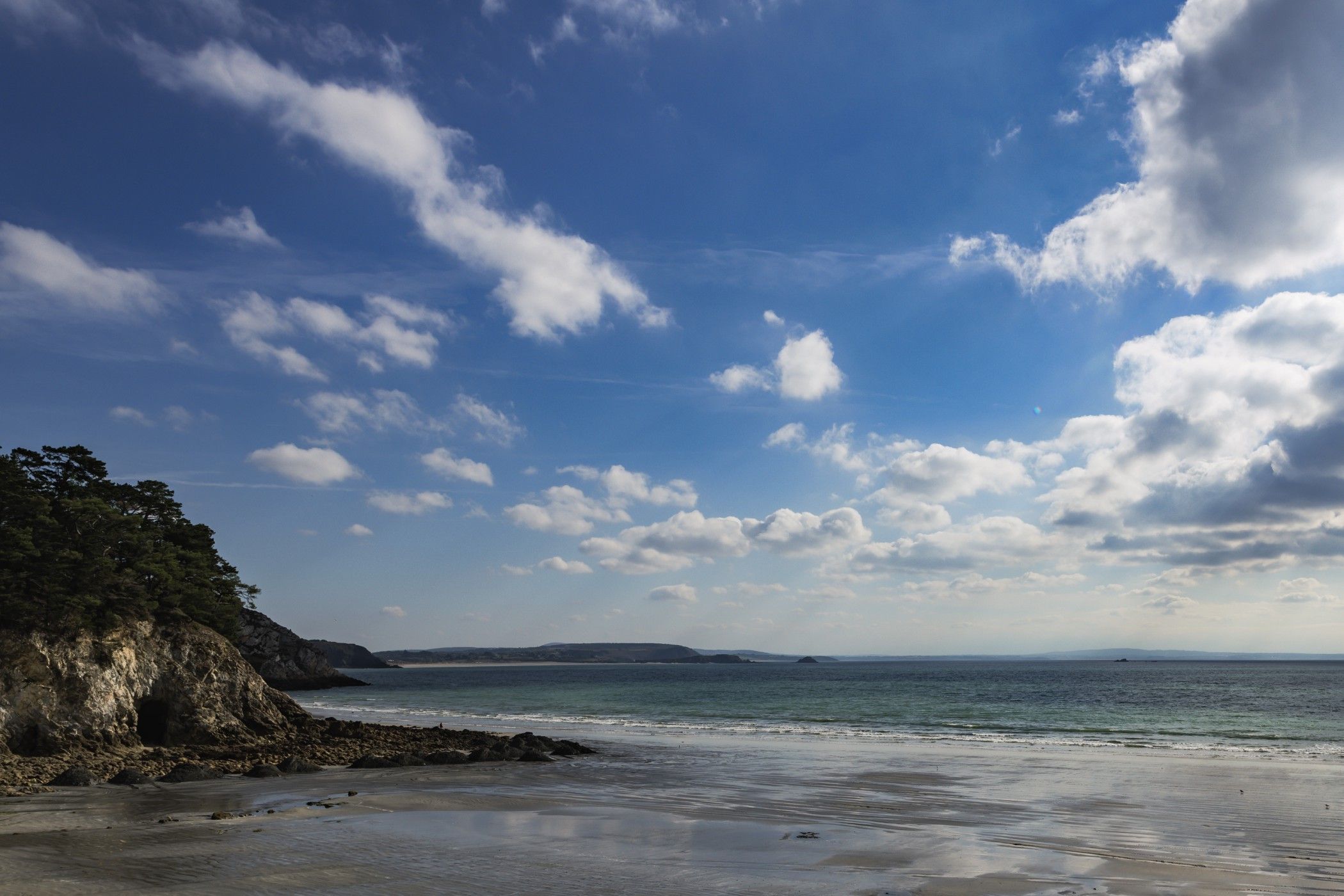 Platja amb un penya-segat rocos, mar tranquil i cel amb algunes boires.