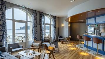 Un hall d’hôtel moderne doté de grandes fenêtres, d’un bar et de différents espaces aménagés pour s’asseoir.