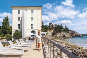 Un hôtel situé sur la côte, doté d’une piscine, de chaises longues et offrant une vue sur la mer.