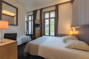 Quarto de hotel com duas camas, uma escrivaninha, uma cadeira, televisão e janelas amplas.