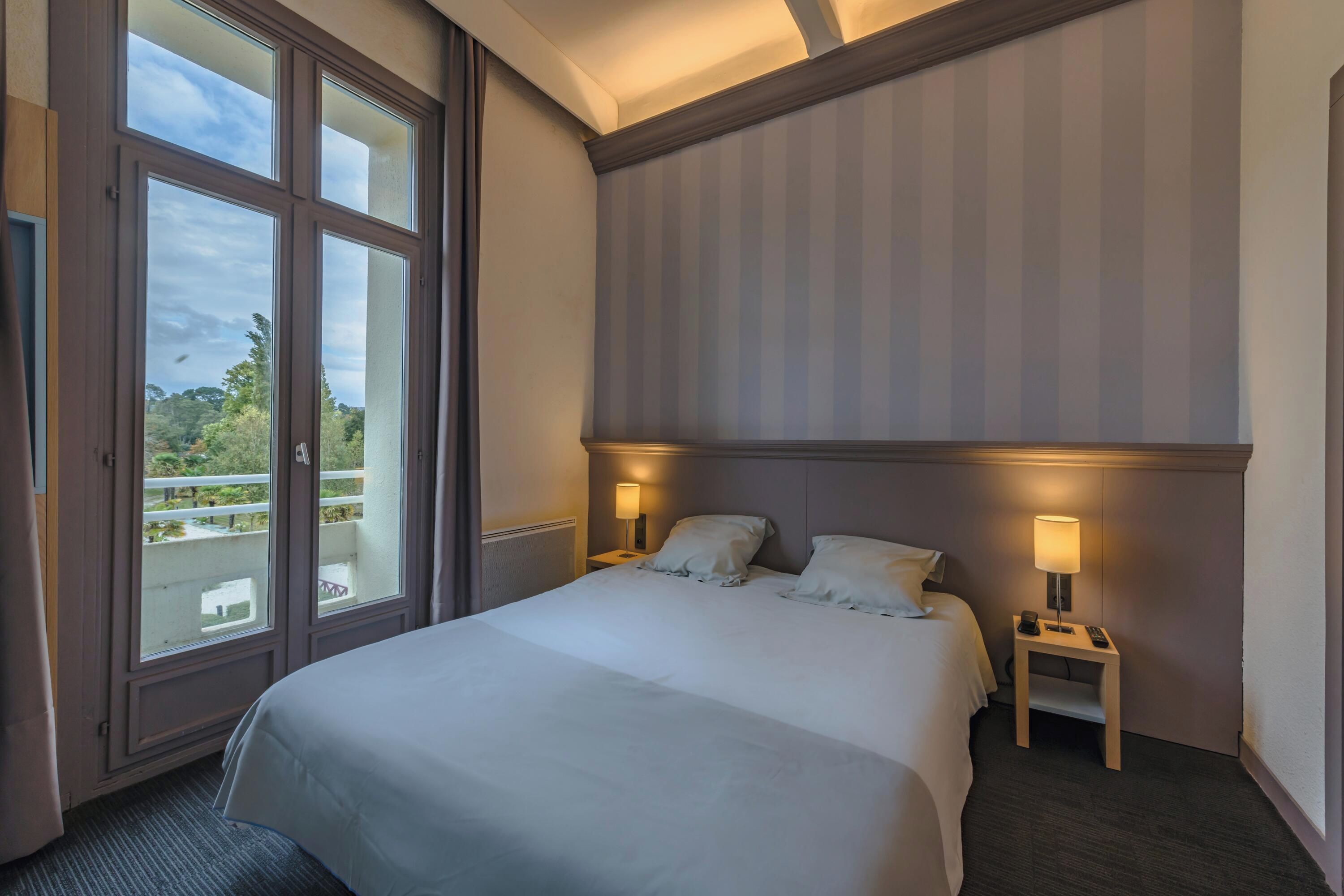Une chambre d’hôtel avec un grand lit, deux tables de chevet avec des lampes, une vue sur la verdure et un mur à rayures.