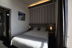 Hotellihuone, jossa on suuri sänky, kaksi yöpöytää valaisimineen, taulu seinällä ja ovi, joka johtaa toiseen huoneeseen.
