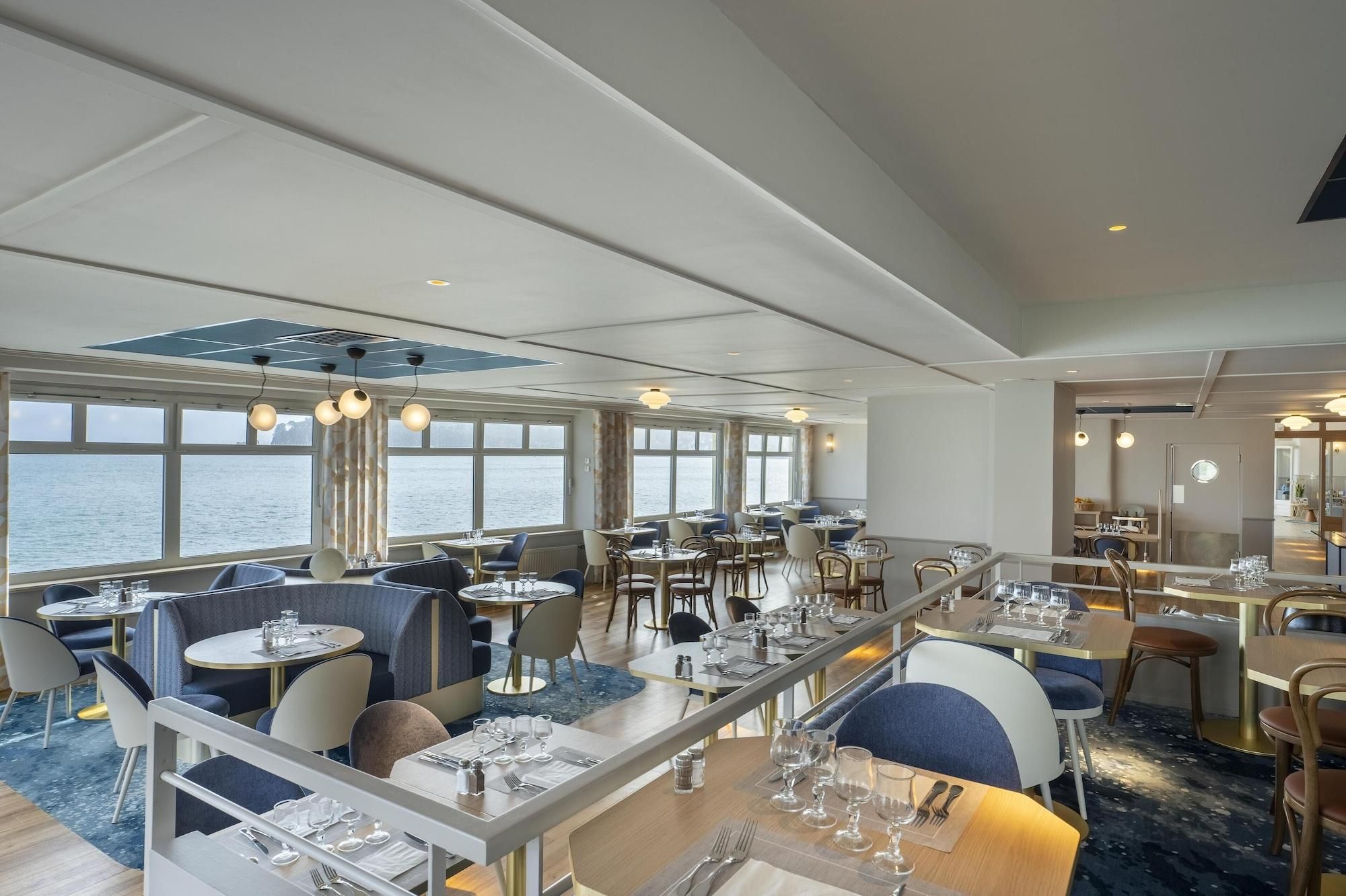 A restaurant with tables and chairs, large windows with a view of the sea, and hanging pendant lights.