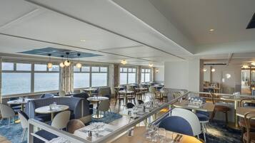 A restaurant with tables and chairs, large windows with a view of the sea, and hanging pendant lights.