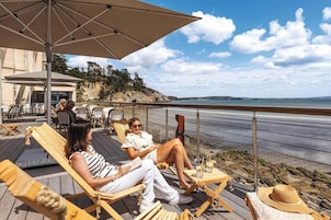 Una terraza con sillas de madera y un paraguas grande, con vista a una playa de costa rocosa y cielo despejado.