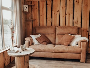 Ein gemütliches Zimmer mit einer Holzwand, einem beigefarbenen Sofa mit Kissen, einem kleinen runden Holztisch und einer Vase mit Trockenblumen.