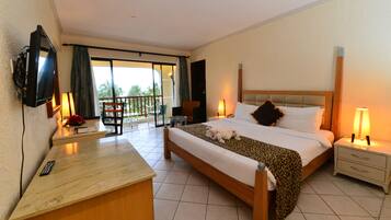 A hotel room with a bed, a desk with a TV, a chair, a nightstand, and a balcony with a view of palm trees.