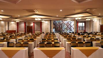 Un salón de conferencias con filas de mesas y sillas, ventanas amplias y una cortina roja.