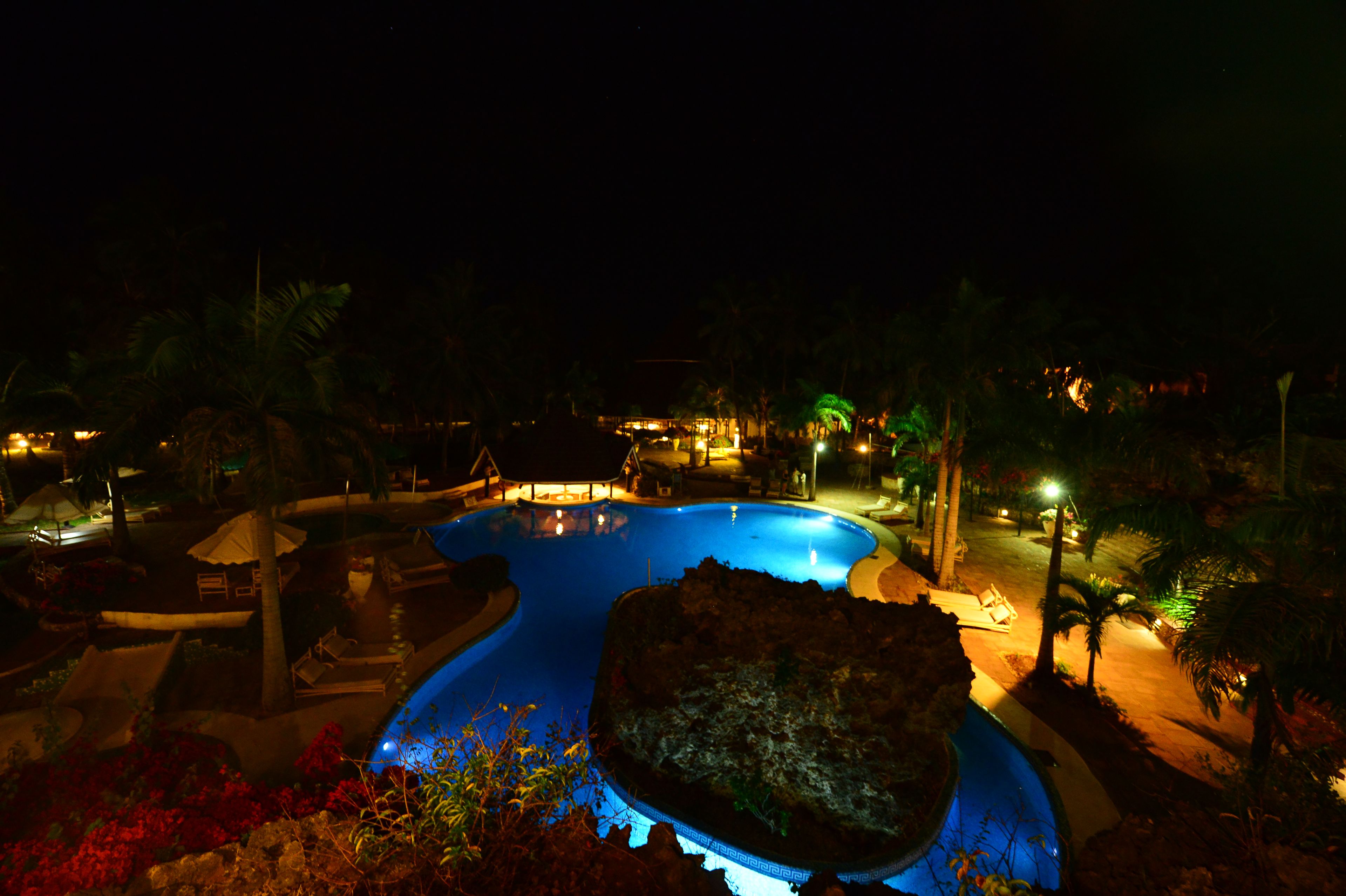 A nighttime view of a resort with a large swimming pool, palm trees, and outdoor seating areas.
