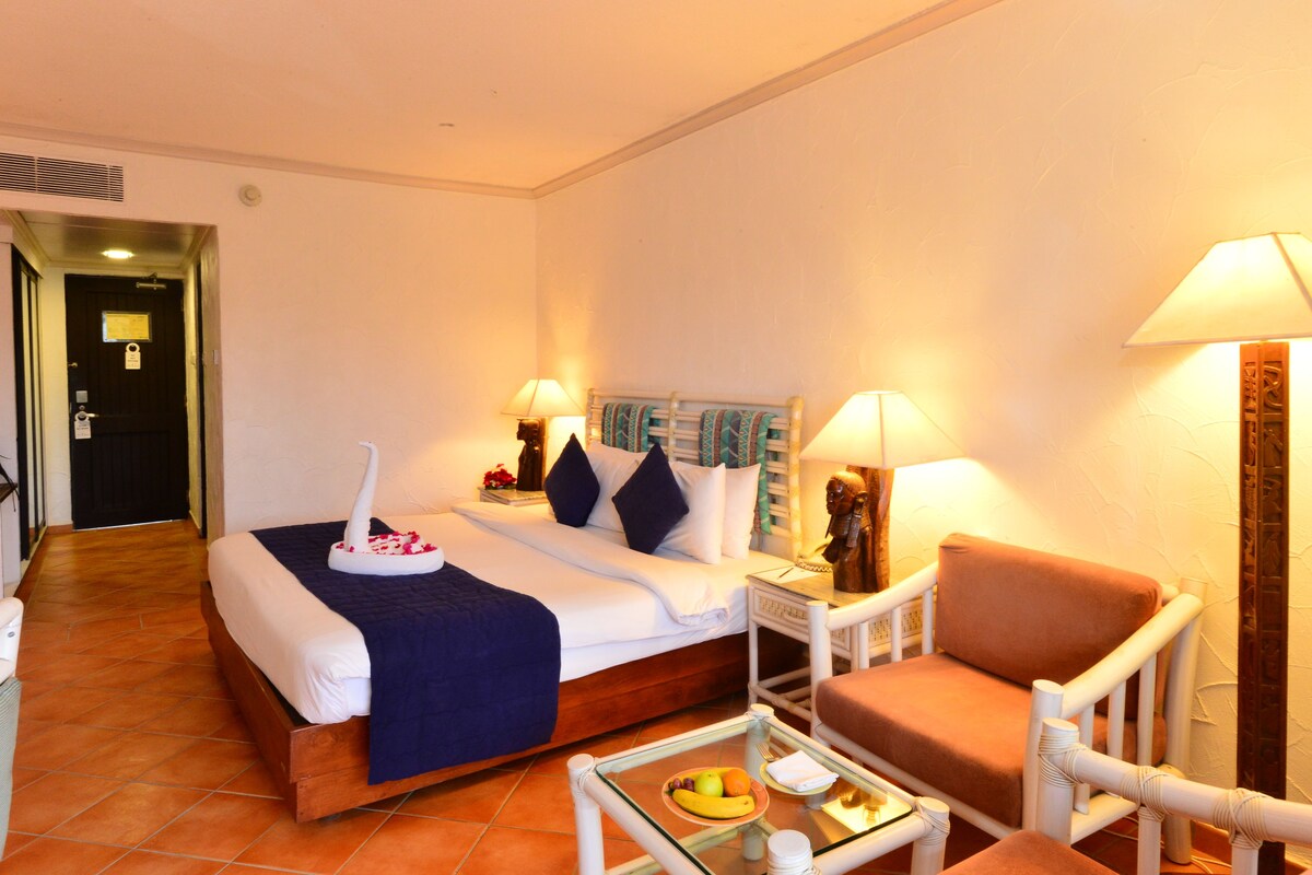 A hotel room with a bed, a chair, a small table with fruit, and two lamps.