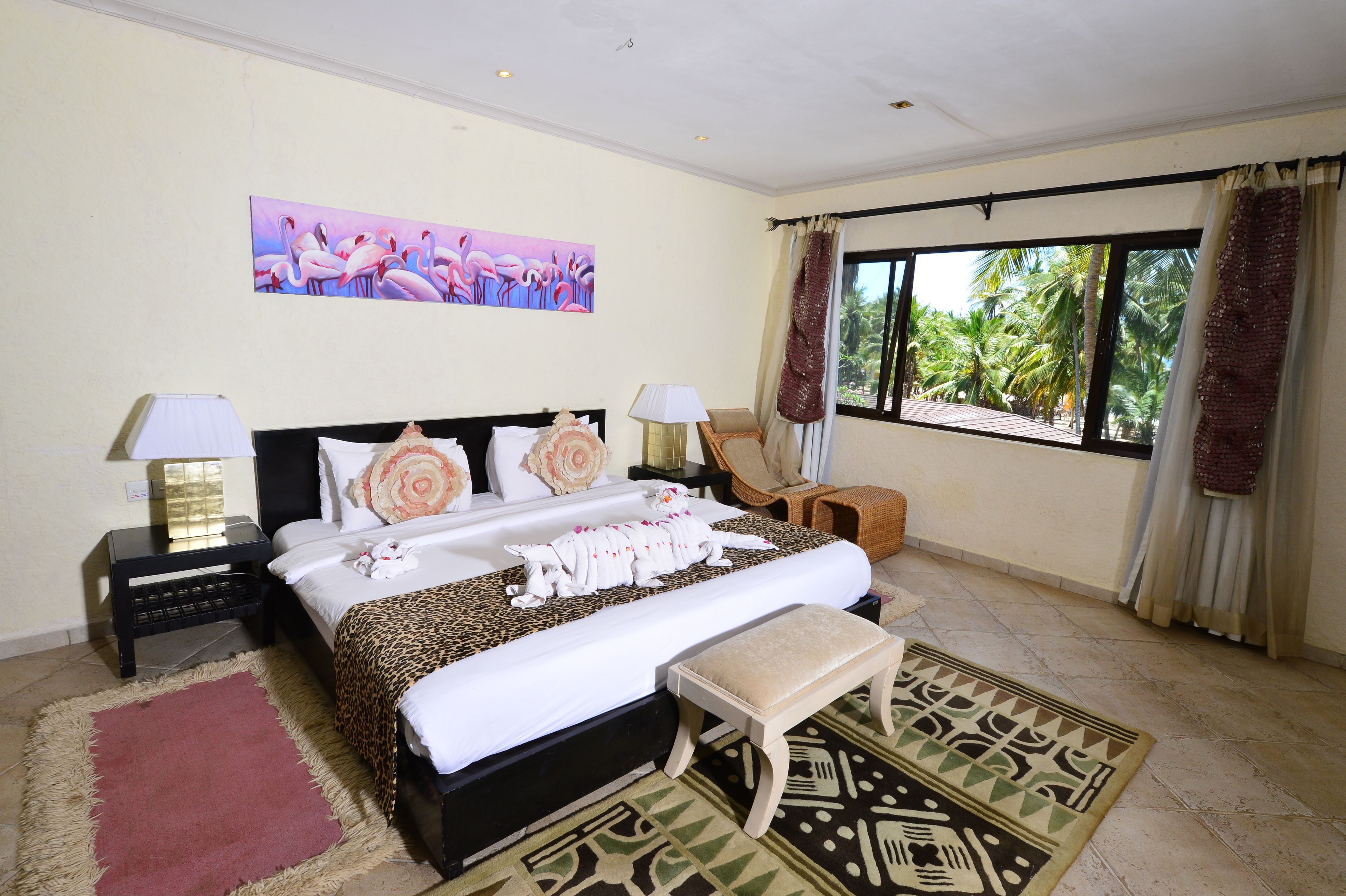 A hotel room with a large bed, two bedside tables, a nightstand, a bench, a patterned rug, a painting of flamingos, and a window with curtains.