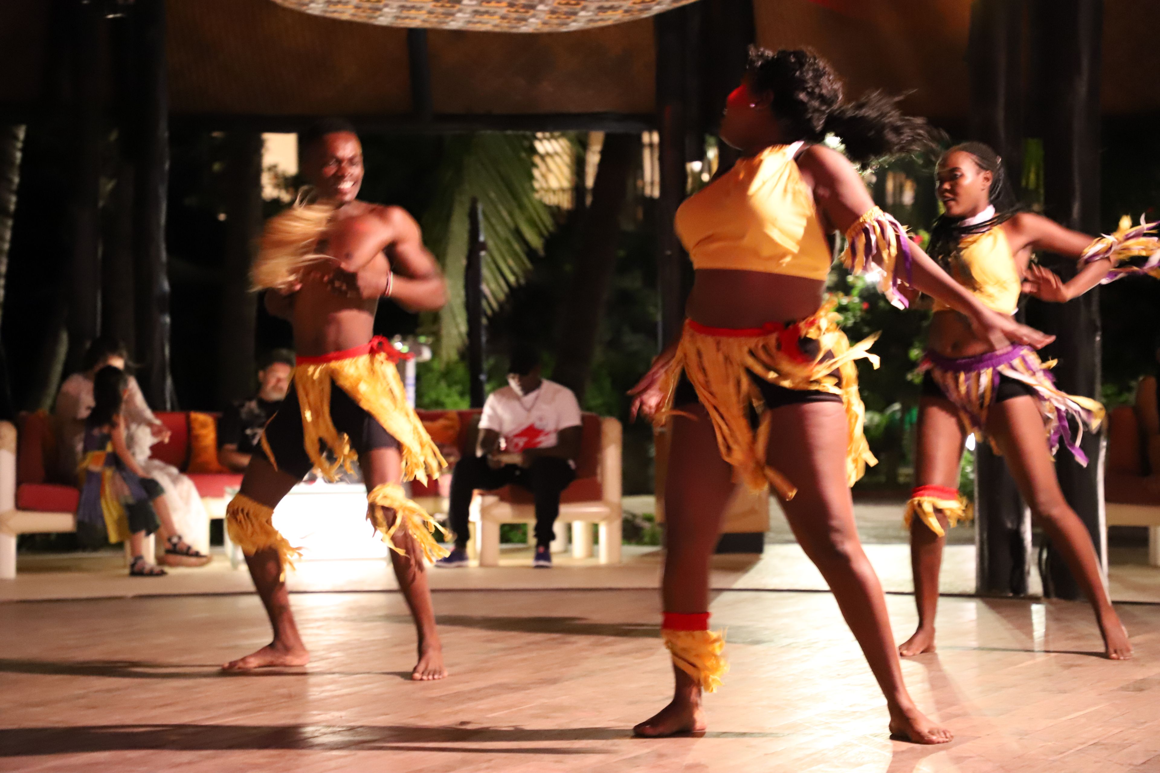 A group of people performing a dance in an outdoor setting with a tiled floor and a bench in the background.