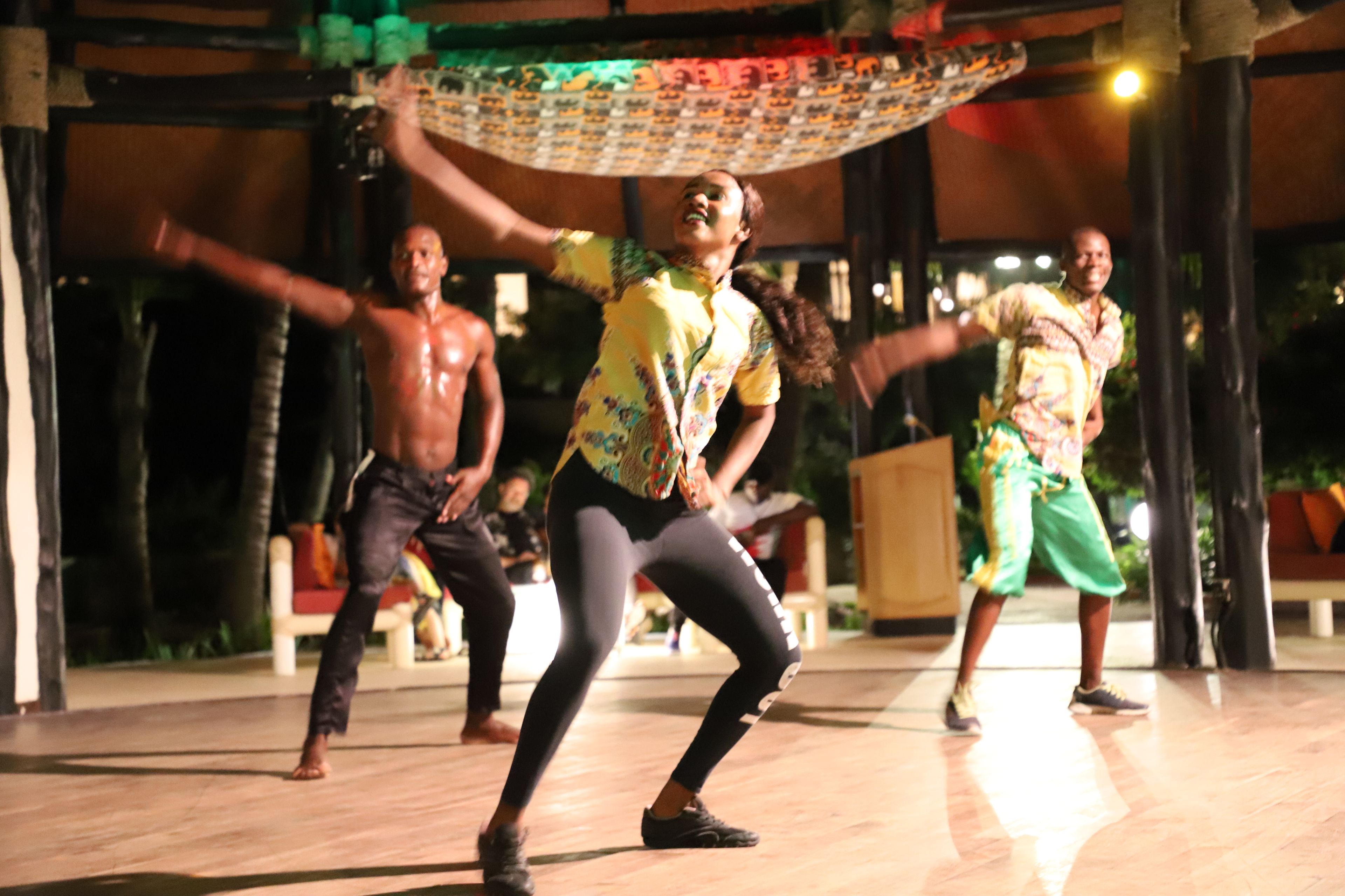 A group of people performing a dance in an outdoor setting with a wooden floor and a covered area.