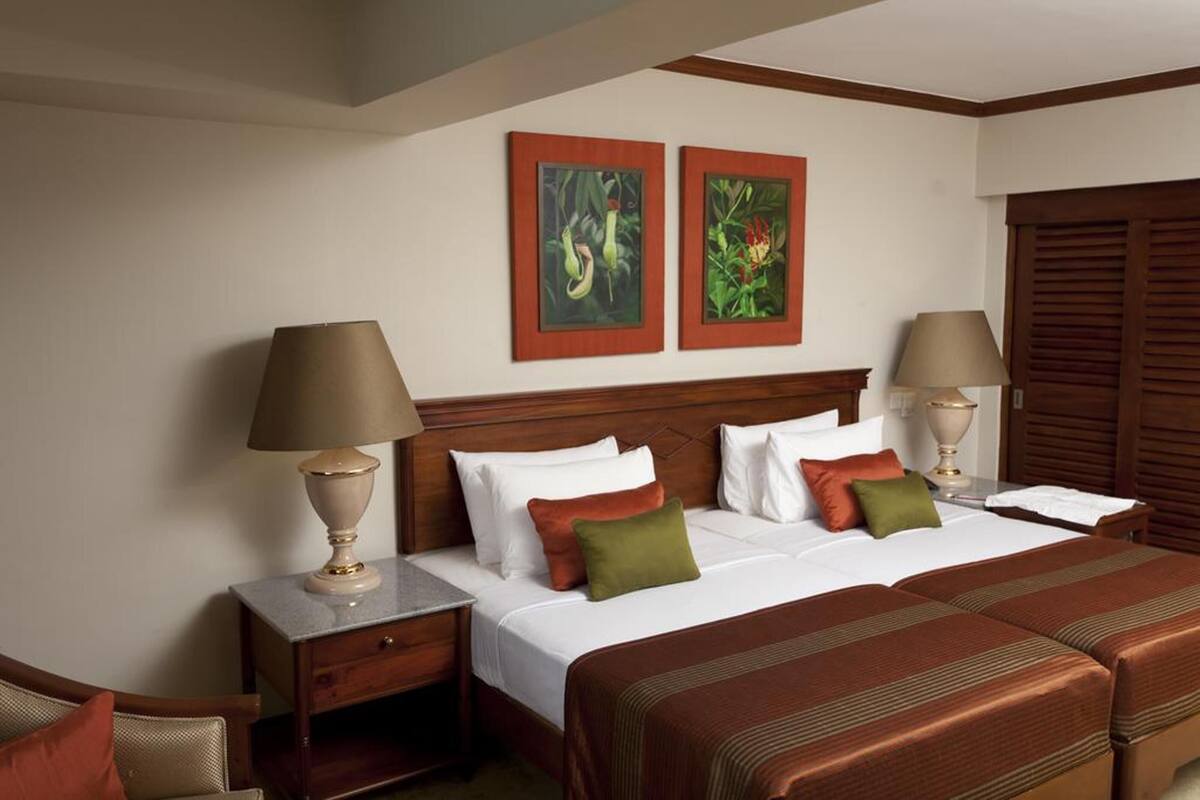 Quarto de hotel com uma cama de casal, duas mesas de cabeceira, um painel de cabeceira de madeira, uma escrivaninha e duas luminárias fixadas na parede.