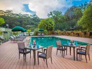 Un espace aménagé au bord de la piscine, avec des chaises longues et des tables, entouré de verdure et d’arbres.