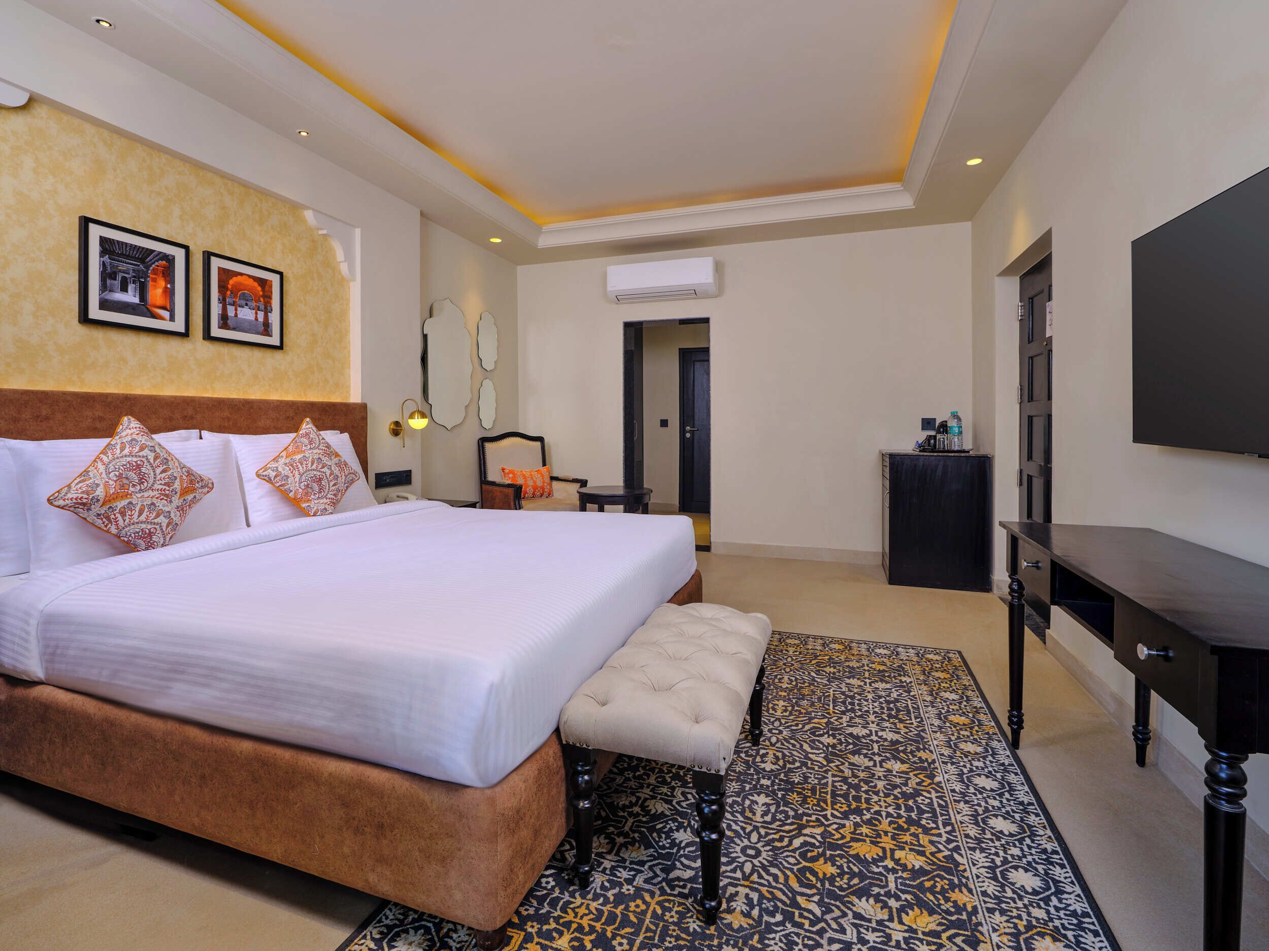 Quarto de hotel com uma cama grande, um banco, uma televisão e um tapete estampado.