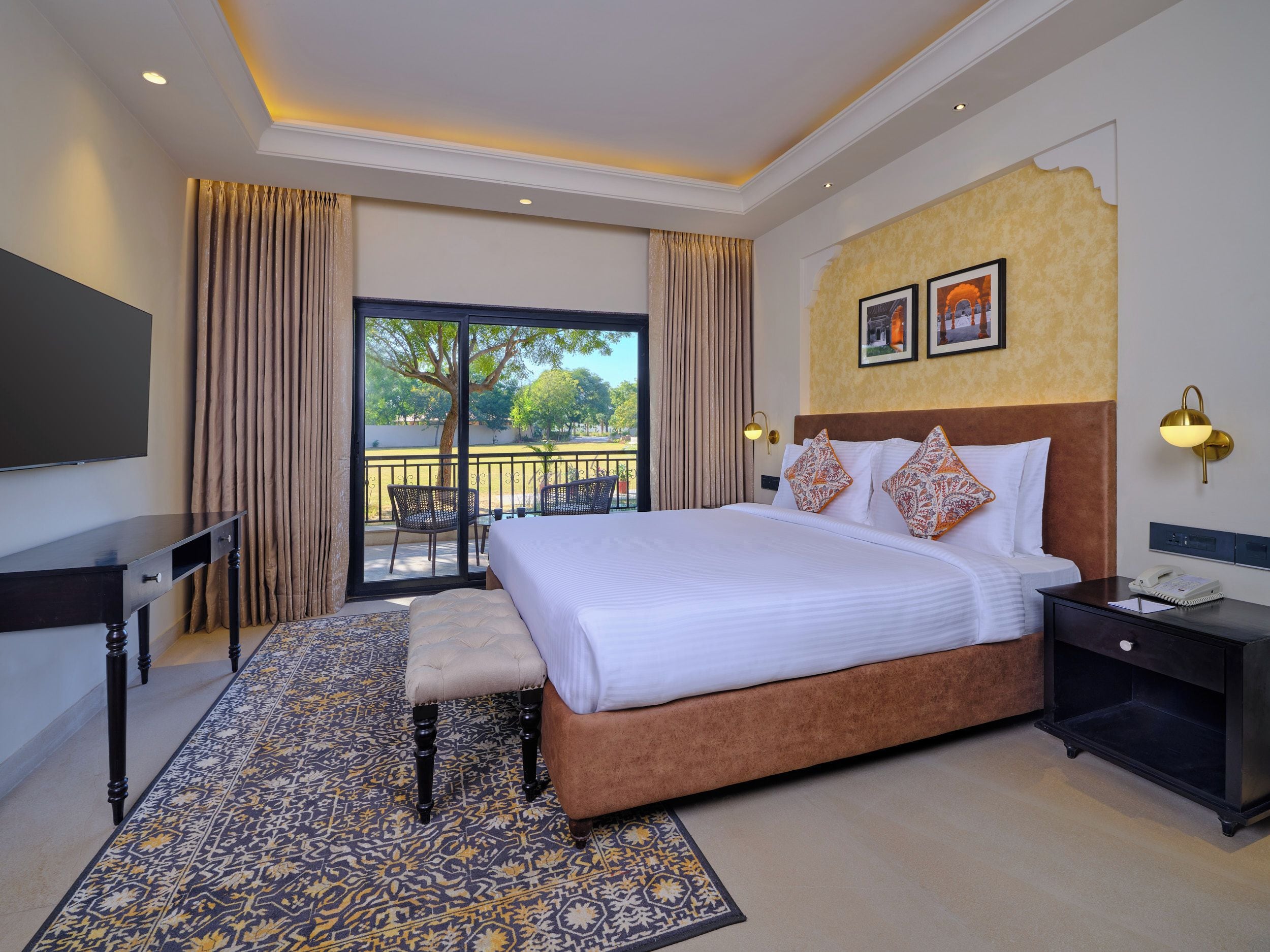 A hotel room with a large bed, a desk, a chair, a small table, a telephone, and a balcony with a view of trees.