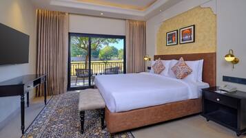 A hotel room with a large bed, a desk, a chair, a small table, a telephone, and a balcony with a view of trees.