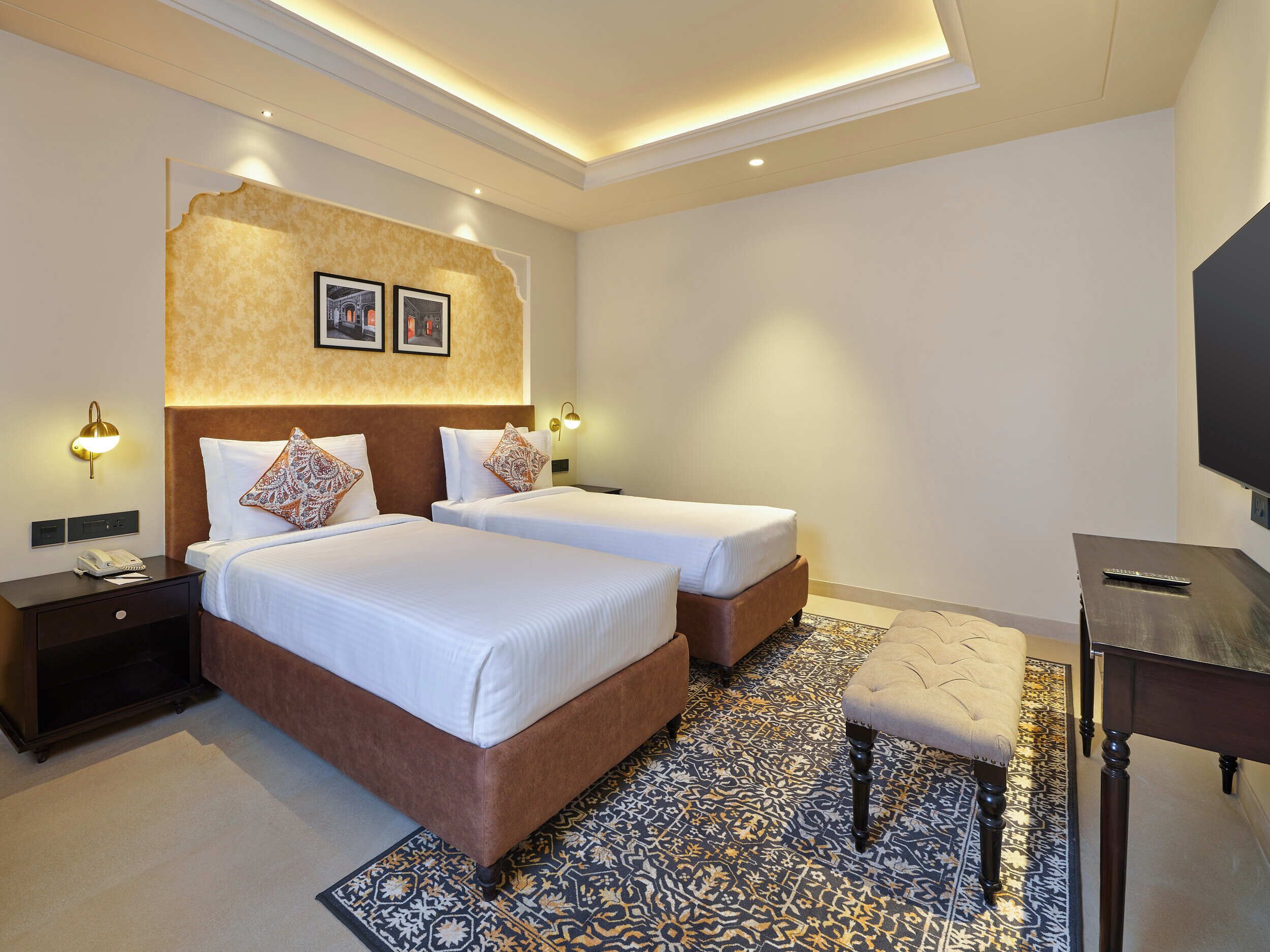Quarto de hotel com duas camas, uma escrivaninha, uma televisão e um tapete estampado.