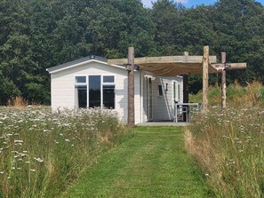 Eine kleine weiße Holzhütte mit einer überdachten Veranda, umgeben von einem grasbewachsenen Feld und dichtem Wald.