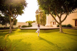 A courtyard with a person performing a yoga pose, surrounded by trees and a building with a dome structure in the background.