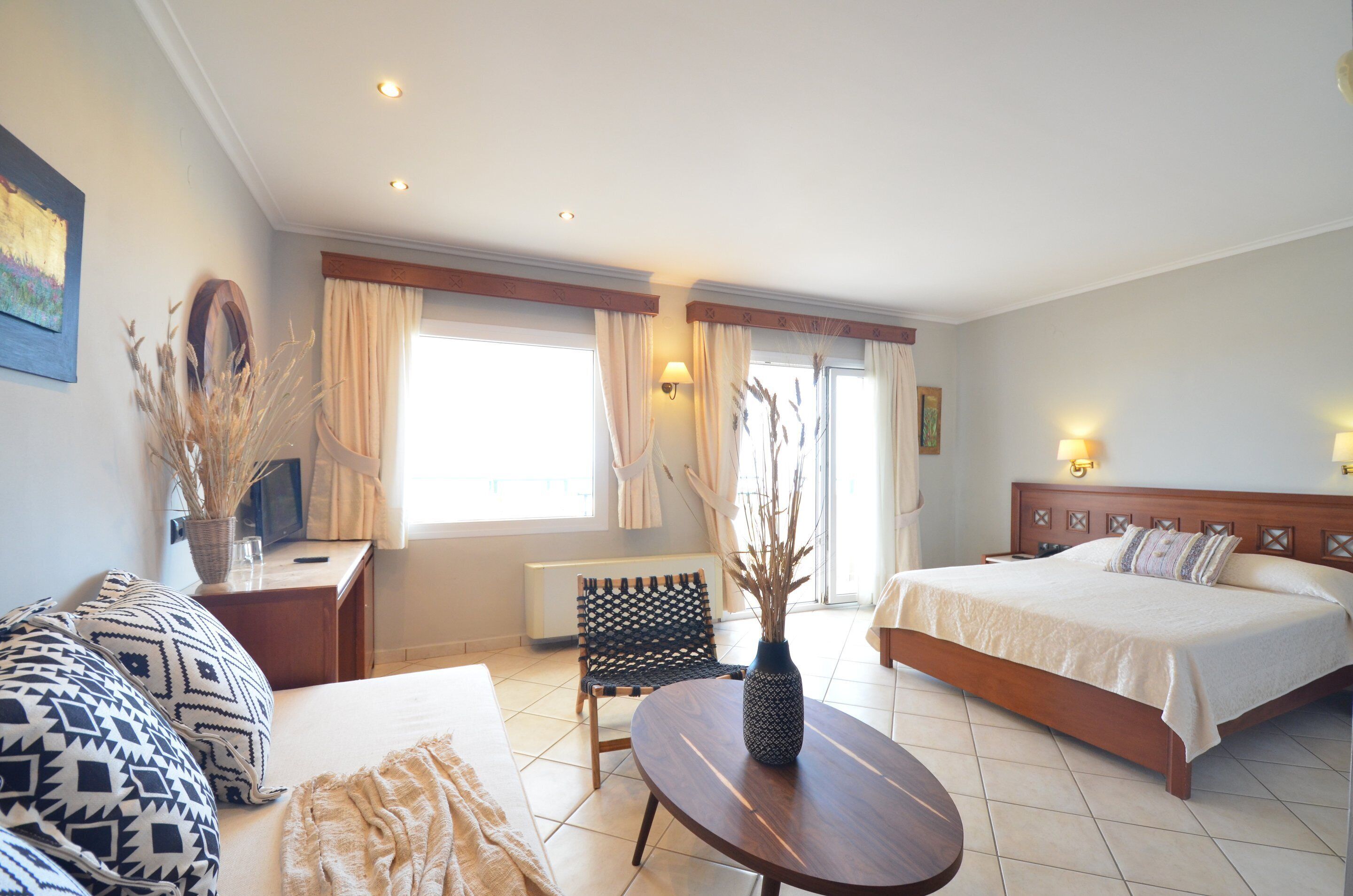 A hotel room with a bed, a sofa, a small table, a chair, a vase with dried plants, and a window with curtains.