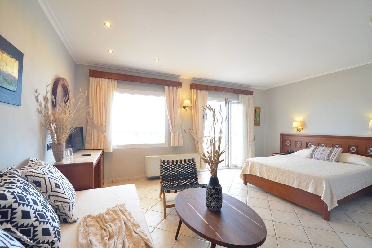 A hotel room with a bed, a sofa, a small table, a chair, a vase with dried plants, and a window with curtains.