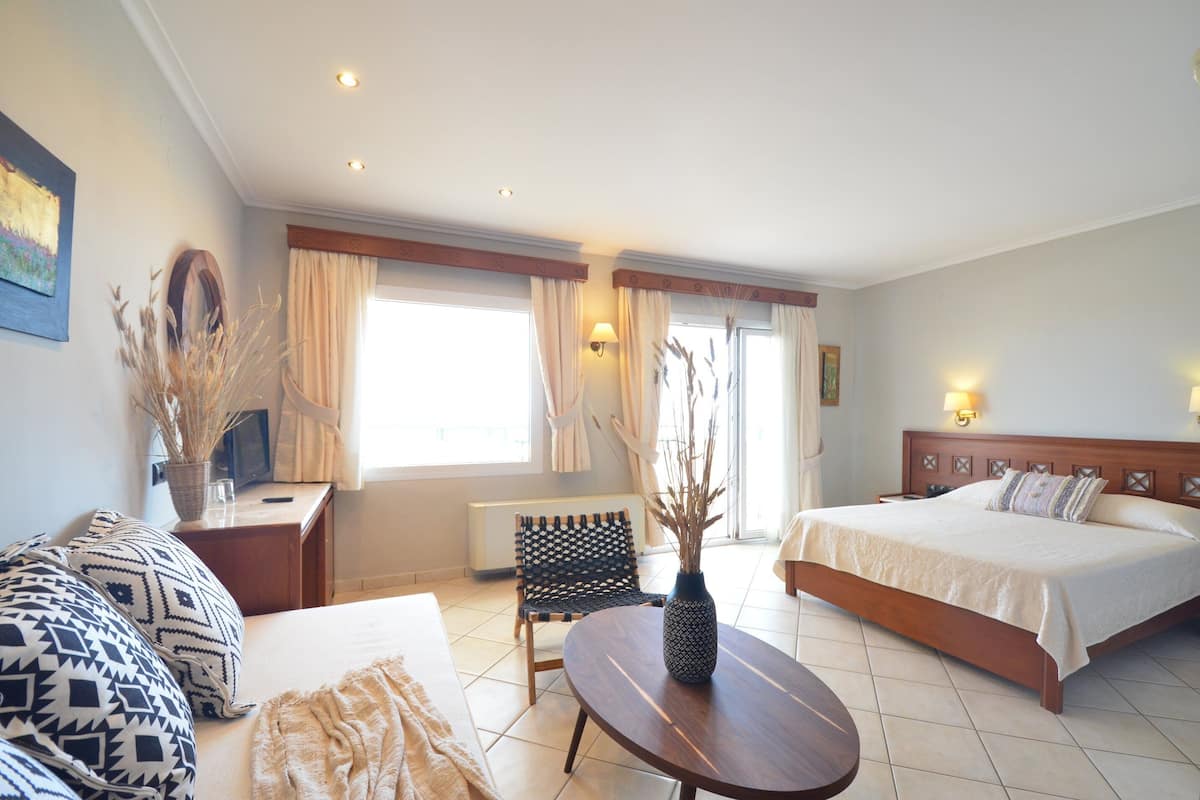 A hotel room with a bed, a sofa, a small table, a chair, a vase with dried plants, and a window with curtains.