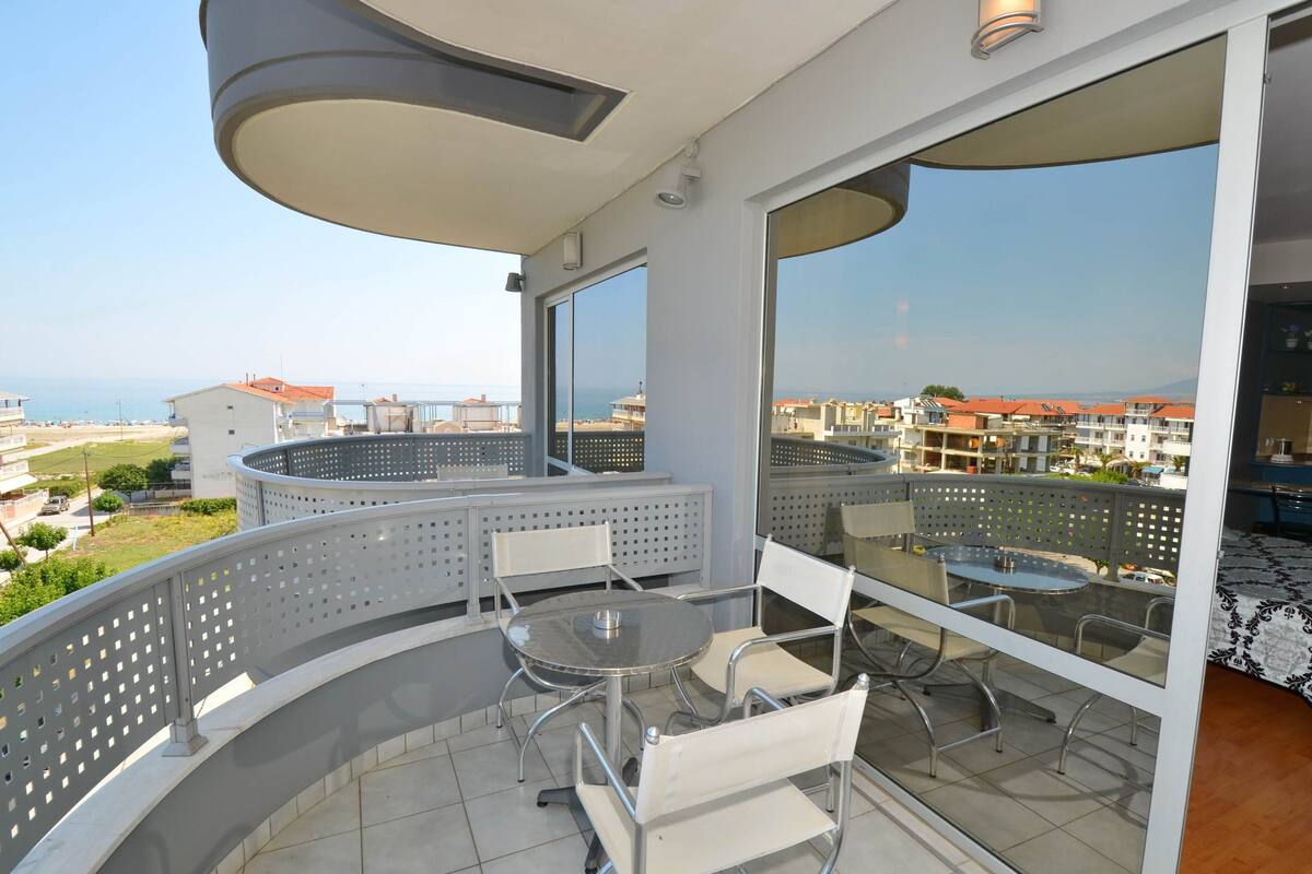 A balcony with a round table and chairs, large windows, and a view of the sea and buildings.