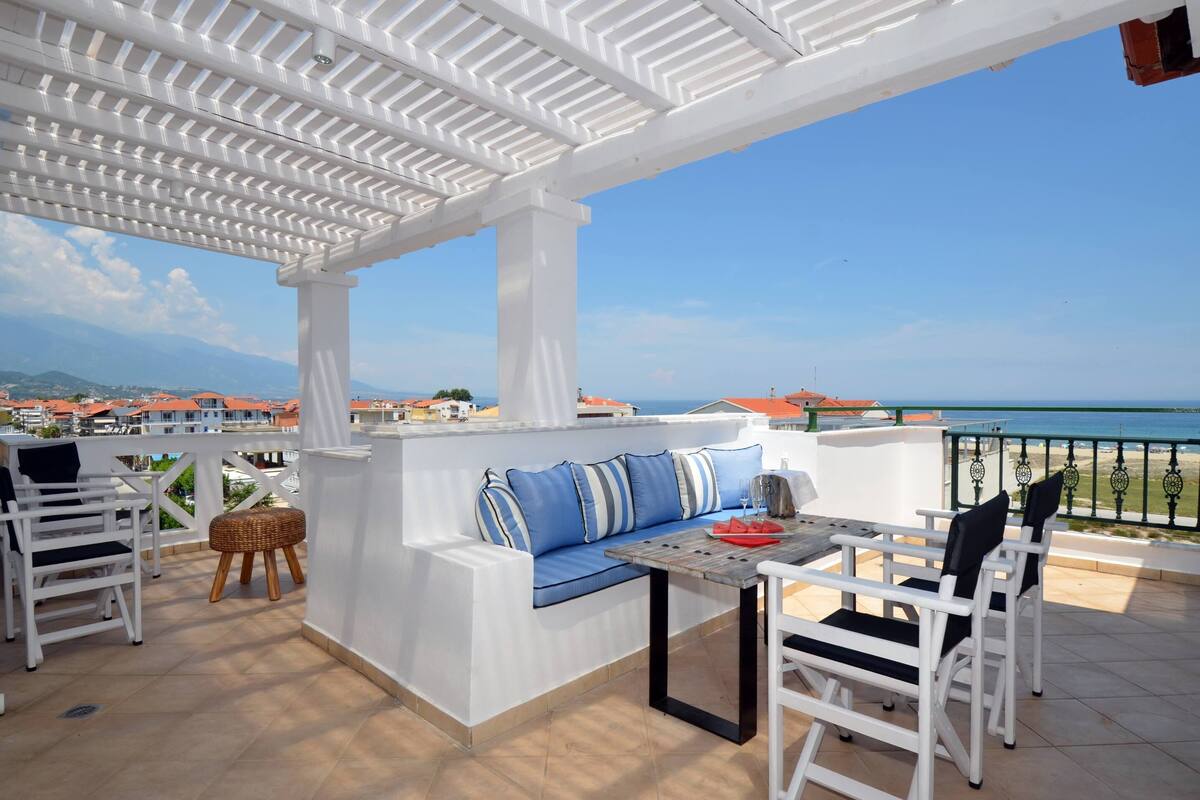 A covered outdoor seating area with a table and chairs, overlooking a coastal view.