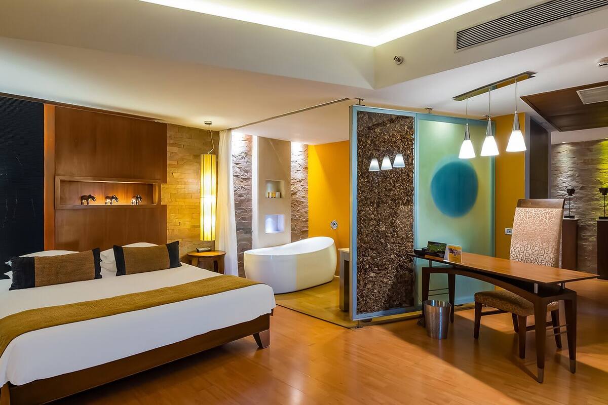 Quarto de hotel moderno com cama grande, banheiro separado, área de refeições e parede de destaque em pedra.