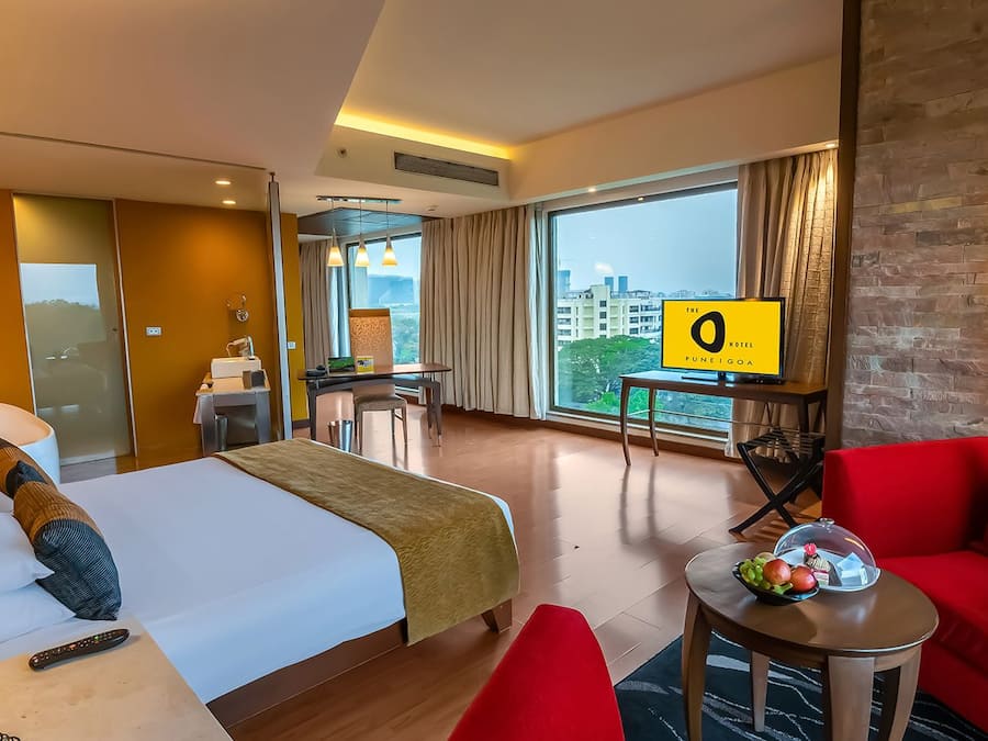Een hotelkamer met een bed, een rode bank, een klein tafeltje met fruit, een televisie en uitzicht op de stad.