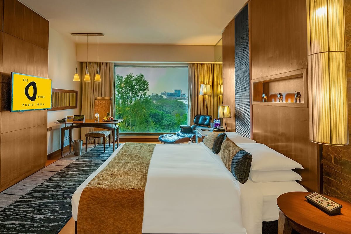 Habitación de hotel con una cama grande, un escritorio, una silla, una lámpara y vistas a la ciudad.