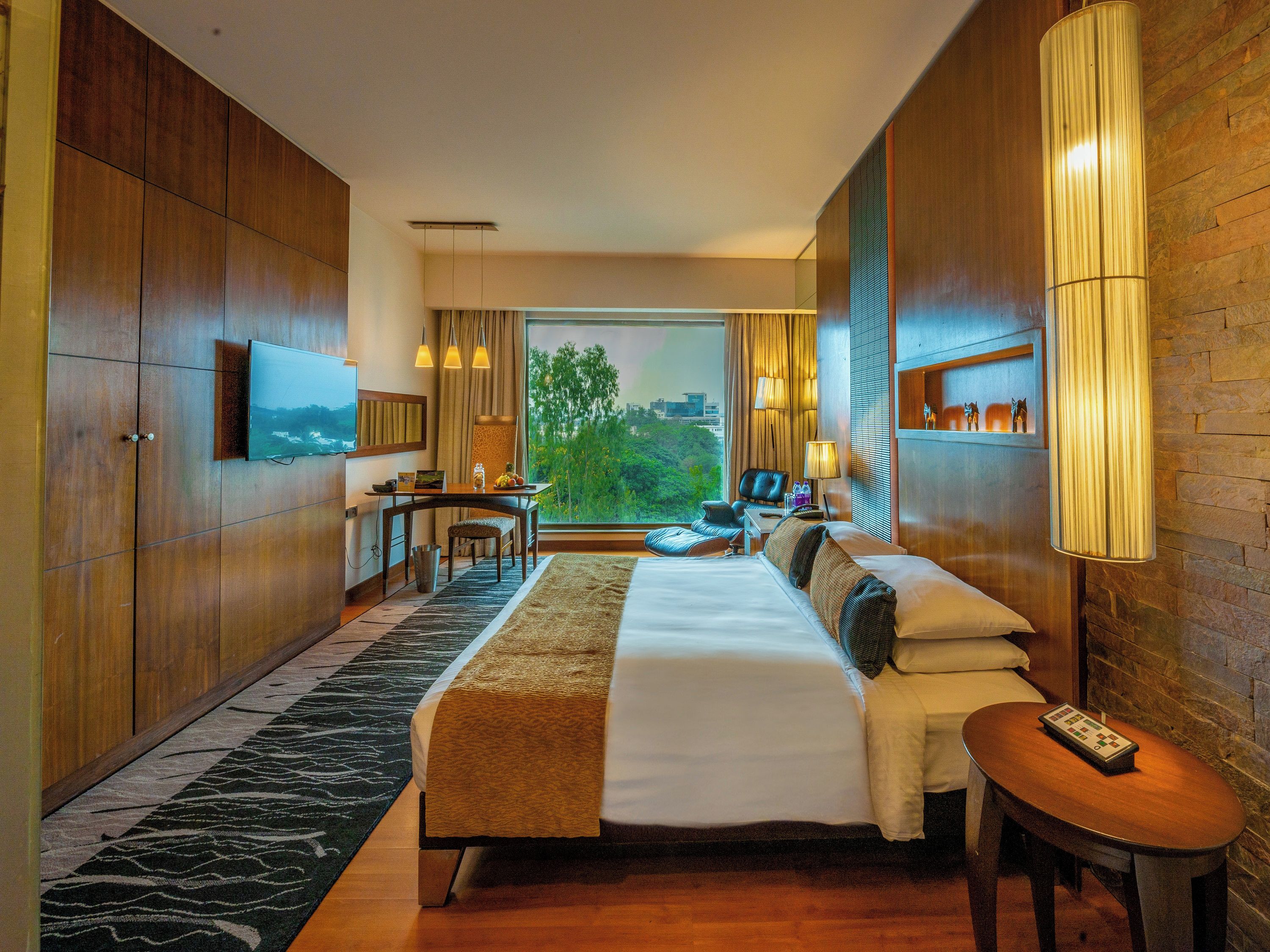A hotel room with a bed, a desk, a chair, a TV, a window with a city view, and a lamp.