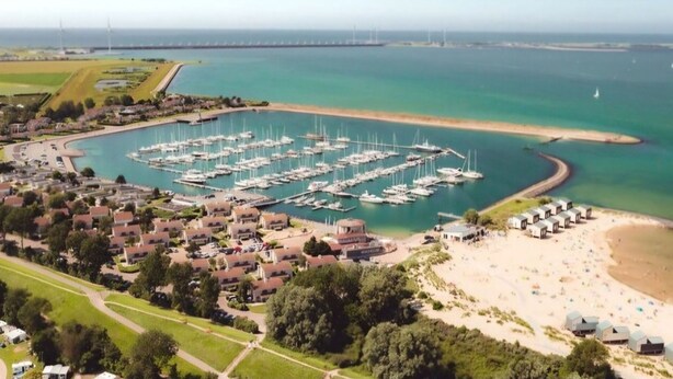 Luftaufnahme einer Marina mit zahlreichen angestuften Booten, einem Sandstrand mit Strandkörben und einer Wohngegend mit Häusern und Grünflächen.
