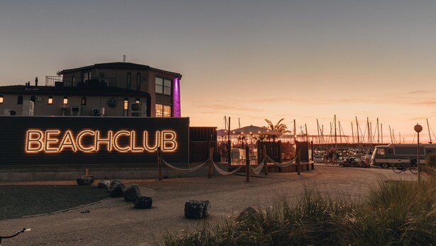 A beach club with a neon sign, a building with a balcony, and a marina in the background. - RBR 1654 - Beach Resort Kamperland by Interhome (Kamperland)