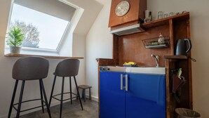Een kleine keuken met blauwe kastjes, een houten aanrecht, een klein krukje, een plant in een pot en een raam.