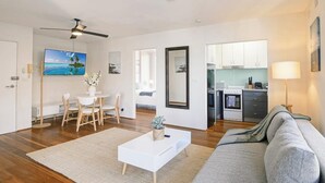 A modern living room with a white coffee table, grey sofa, wooden flooring, a dining area with a white table and chairs, a kitchen with white cabinets, and a ceiling fan. - KozyGuru | Manly | Prime Manly Coastal Hub (Manly)