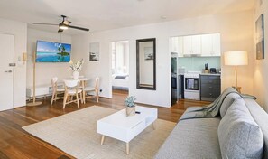 Ein modernes Wohnzimmer mit einem weißen Couchtisch, einem grauen Sofa, Holzböden, einem Essbereich mit weißem Tisch und Stühlen, einer Küche mit weißen Schränken und einem Deckenventilator.