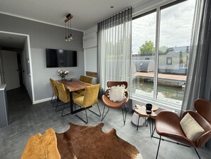 A modern living room with a dining area, a TV mounted on the wall, and a view of a water body and buildings outside. - Maasterp - Woonboot 2 by Interhome (Ohe en Laak)