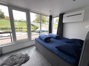 A bedroom with a large window, a bed with blue bedding, a grey rug, and a white air conditioning unit. - Maasterp - Woonboot 2 by Interhome (Ohe en Laak)