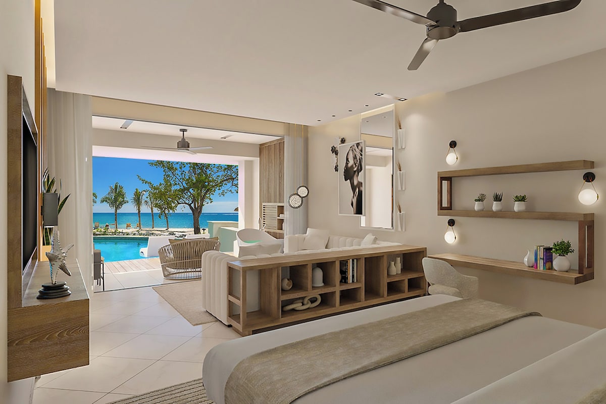 A modern bedroom with a large bed, a ceiling fan, a wooden nightstand, a bedside table with a lamp, a large mirror, and a view of a pool and beach.