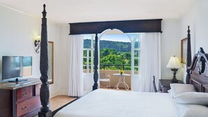 A hotel room with a four-poster bed, a television, a small table with a chair, and a balcony with a view of greenery.
