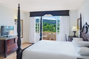 A hotel room with a four-poster bed, a television, a small table with a chair, and a balcony with a view of greenery. - Sandals Dunn's River Falls (Ocho Rios)