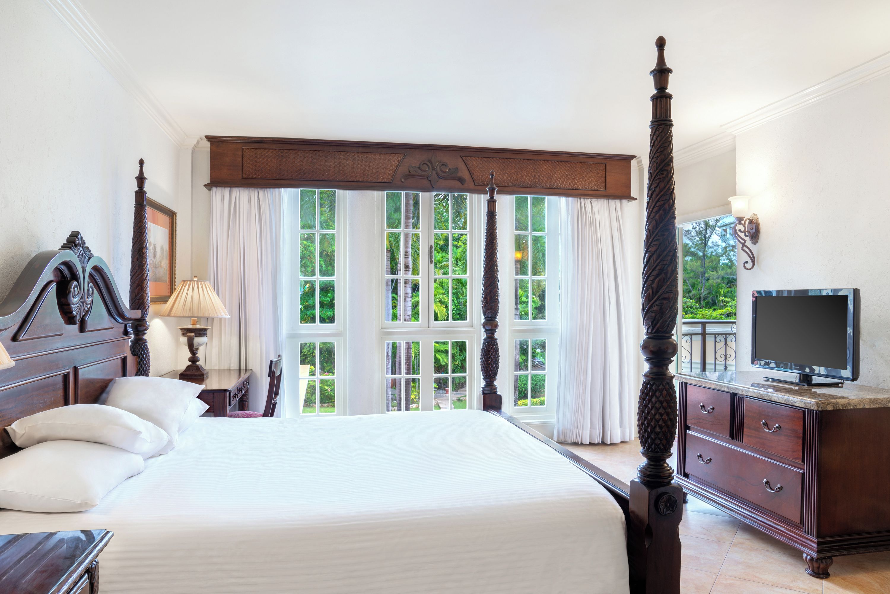 A spacious bedroom with a large bed, a television, and a view of greenery through the windows.