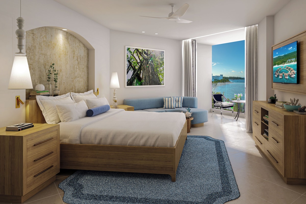 A modern hotel room with a bed, a TV, a balcony with a view of the sea, and a desk with a lamp.