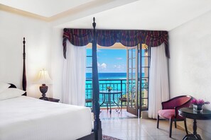 A hotel room with a four-poster bed, a nightstand with a lamp, a chair, a small table with a vase, and a balcony with a view of the ocean. - Sandals Dunn's River Falls (Ocho Rios)