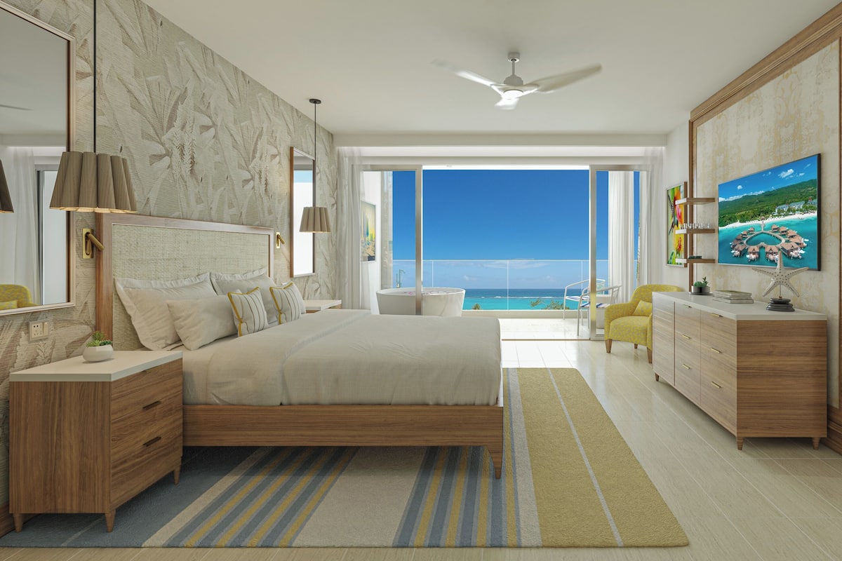 A modern bedroom with a large bed, a TV, a dresser, and a view of the ocean.