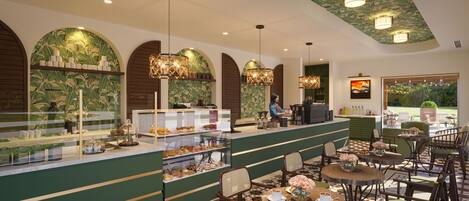 A café with a counter displaying pastries, seating area with round tables and chairs, and a view of greenery outside.